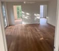 Empty room with light wooden flooring, white walls, and large windows allowing natural light. The room has two open doorways leading to other spaces.