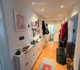 A narrow hallway with wooden flooring, white walls, and several doors leading to other rooms. There are storage cabinets, a coat rack with jackets, and a colorful runner rug.