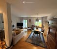 A well-lit living and dining area with wooden flooring, a sofa, coffee table, dining table with chairs, television, fan, and decorative items.