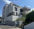 Modern two-story house with white facade, large windows, and multiple balconies, surrounded by greenery and a concrete driveway.