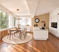 Modern kitchen with white cabinets, wooden dining table, six chairs, and a kitchen island with sink and oven. Large windows with sheer curtains, pendant light, and a round rug.