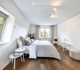 Modern, minimalist bedroom with a large bed, nightstands, table lamps, a chair, a stool, and a vase with flowers. The room features wooden flooring and large windows with curtains.