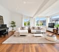 Bright living room with white walls, wooden floors, large windows, and ample natural light. Features a white sectional sofa, coffee table, ottoman, piano, and television. Decor includes plants, a throw blanket, and a chessboard on a round table.
