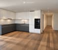 Modern hallway with wooden flooring, white walls, recessed lighting, and a view into a contemporary kitchen with white and grey cabinets, a sink, and built-in ovens.