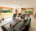 Bright living room with large windows, leather sofa, coffee table, armchairs, ottoman, dining area with table and chairs, and decorative plants.