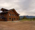 Two-story wooden house with multiple windows and a garage, surrounded by a grassy field with cows grazing in the background and mountains visible in the distance.