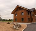 Two-story wooden house with multiple windows and a sloped roof, surrounded by a paved area and scattered rocks.