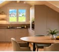 Modern kitchen with light wood cabinetry, stainless steel appliances, and a large window offering a scenic view of a green field and mountains. A round dining table with chairs is placed near the window, accompanied by a bowl of fruit and a vase with plan
