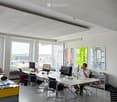 Modern office space with large windows, multiple desks, chairs, computers, and a person working. Posters and artwork are displayed on the walls.