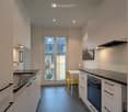 Modern kitchen with white cabinets, black countertops, stainless steel sink, oven, and a small dining area with a yellow chair and white table. A large window offers a view of the street.