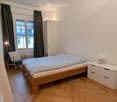 Cozy bedroom with a wooden bed, a nightstand with a lamp, a chair, and a wardrobe. The room features wooden flooring and large windows with dark curtains.