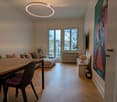 Modern living room with a sofa, dining table, and chairs, featuring large windows with a city view, a colorful painting on the wall, and a round pendant light.
