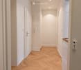 A narrow hallway with light wooden herringbone flooring, white walls, and recessed ceiling lights. Doors are visible on both sides, and a small section of a kitchen area is visible on the right.