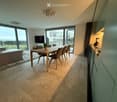 Modern open-plan living and dining area with large windows offering a scenic view, featuring a sofa, dining table with chairs, television, and a kitchen area with cabinets and appliances.