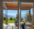 Bright room with large glass doors opening to a patio, featuring a view of a lake and mountains, with a vase and a basket visible inside.