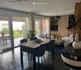 Modern dining and living area with a large wooden dining table, chairs, a sofa, coffee table, and decorative vases. Large windows offer a view of the garden and sea.