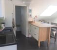 Modern kitchen with a black induction stove, white cabinets, a wooden kitchen island with drawers, and a small table with chairs. A potted plant and a lamp are visible, along with a doorway leading to a bathroom.