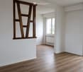 Empty room with light wooden flooring, white walls, and a large window with a view. A wooden shelf is mounted on the wall, and there is a sliding glass door leading to another space.