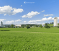 Open green field with scattered trees and distant buildings under a partly cloudy sky.