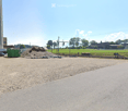 Gravel driveway leading to a construction area with a large pile of dirt, a truck, and a green container. A tall concrete silo is visible in the background, with a grassy field and scattered trees beyond.