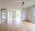 Bright, empty room with light wooden flooring, large glass sliding doors leading to a balcony, white walls, and modern lighting fixtures.