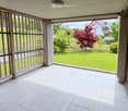 Spacious balcony with white tiled flooring, wooden railings, and large glass panels offering views of a well-maintained garden and surrounding houses.
