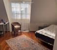 Cozy attic bedroom with wooden floors, a large window with blinds, a bed with patterned bedding, a wicker chair, a desk, and a nightstand.
