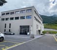 Modern two-story building with large windows and a garage entrance, surrounded by a parking lot with several cars and a scenic mountain backdrop.