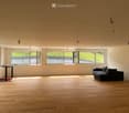 Bright, open living space with wooden flooring, large windows, a black sectional sofa, and a small table with books.