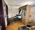 A compact, furnished room with a bed, sofa, desk, chair, wardrobe, and shelves. The room features wooden flooring and large windows with dark curtains.