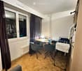 A small, furnished room with a sofa, desk, chairs, and a window with curtains. The room features wooden flooring and a radiator under the window.