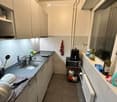 Modern kitchen with white cabinets, stainless steel sink, oven, and various appliances. Countertops hold a few items including a bottle, a cup, and a small basket with fruits.