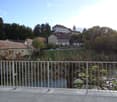 Balcony overlooking a river with a metal railing, trees, and distant buildings visible in the background.