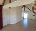 Empty room with wooden flooring, white walls, exposed wooden beams, and two white doors. The room is brightly lit and has a modern, minimalist design.