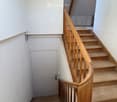 Wooden staircase with a wooden handrail and balusters, leading to an upper level. The walls are white, and there is a door at the end of the hallway.