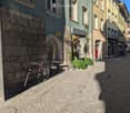 Stone pavement street, multi-story buildings with brick facades, archway entrance, bicycle leaning against a wall, potted plants, streetlight, parked bicycle.