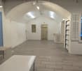 A spacious room with an arch ceiling and gray tile floor. On the left is a door with shelves above it and wall-mounted lights. In the middle is a doorway with an open door to a sink area. On the right, there are white shutters and another doorway. A white