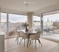 Modern dining room with light wood flooring, large windows, and a round dining table with six chairs. A decorative vase with greenery sits on the table, and a balcony with outdoor furniture is visible through the sliding glass doors.
