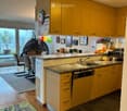 Bright kitchen with yellow cabinets, granite countertop, sink, dishwasher, and various appliances. A small seating area with chairs and a table is visible, along with a colorful rug and balloons hanging from the ceiling.
