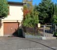 A two-story house with a garage, surrounded by a wooden fence and lush greenery. The driveway leads to the garage, and there is a mailbox and a gate visible.