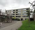 Modern multi-story building with large windows and a playground area in front, featuring wooden play structures and a slide.