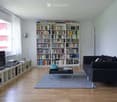 Modern living room with a large bookshelf filled with books, a black sofa, a coffee table, a television on a stand, and a floor lamp near the window.