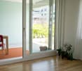 A small, well-lit room with wooden flooring, a red chair, a small table, and several potted plants near the sliding glass door that opens to a garden area.