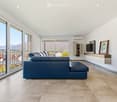 Modern living room with large windows and a balcony overlooking a scenic mountain view. Features a blue sectional sofa, a flat-screen TV on a wall-mounted cabinet, and a framed picture on the wall.