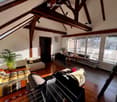 Bright attic living space with wooden beams, large windows, and a mix of seating including a sofa and armchair. Features a small desk area with a chair and a fan, along with a potted plant and a picture frame on the wall.