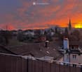 Balcony overlooking a cityscape at sunset, with rooftops, chimneys, and a distant church spire visible against a colorful sky.