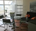 Modern living room with a black sofa, glass table, four chairs, and a shelf with vases. Large windows offer a city view.