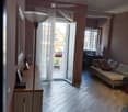 Living room with wooden flooring, large window with sheer curtains, open door leading to a balcony, sofa with guitar, side table with picture frames, and a floor lamp.