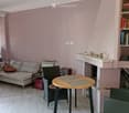A cozy living room with a grey sofa, a round wooden table, wicker chairs, a guitar, and a speaker system. The room features light pink walls, wooden flooring, and built-in shelving with books and decorative items.