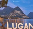 Scenic view of Lake Lugano with illuminated town and mountains in the background, featuring large white letters spelling "MY LUGANO" in the foreground.